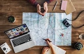 Overhead view of two people planning a trip, map spread on table with laptop, camera, and other travel items.